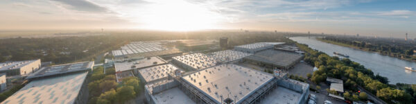 A panoramic view of the Messe Düsseldorf exhibition grounds, located directly on the Rhine, featuring modern exhibition halls with the Düsseldorf cityscape in the background. @ Copyright MD 